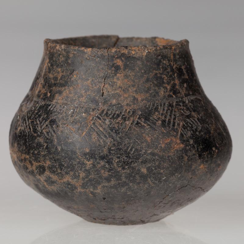 Miniaturowa waza o płaskim dnie, brzuścu baniastym i stożkowatej szyjce. Naczynie zdobione dookolnym żłobkiem i naprzemiennymi grupami kresek.