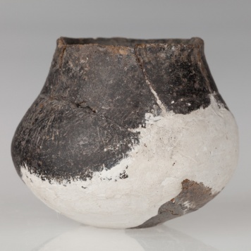 Miniaturowa waza o płaskim dnie, brzuścu baniastym i stożkowatej szyjce. Naczynie zdobione dookolnym żłobkiem i naprzemiennymi grupami kresek.